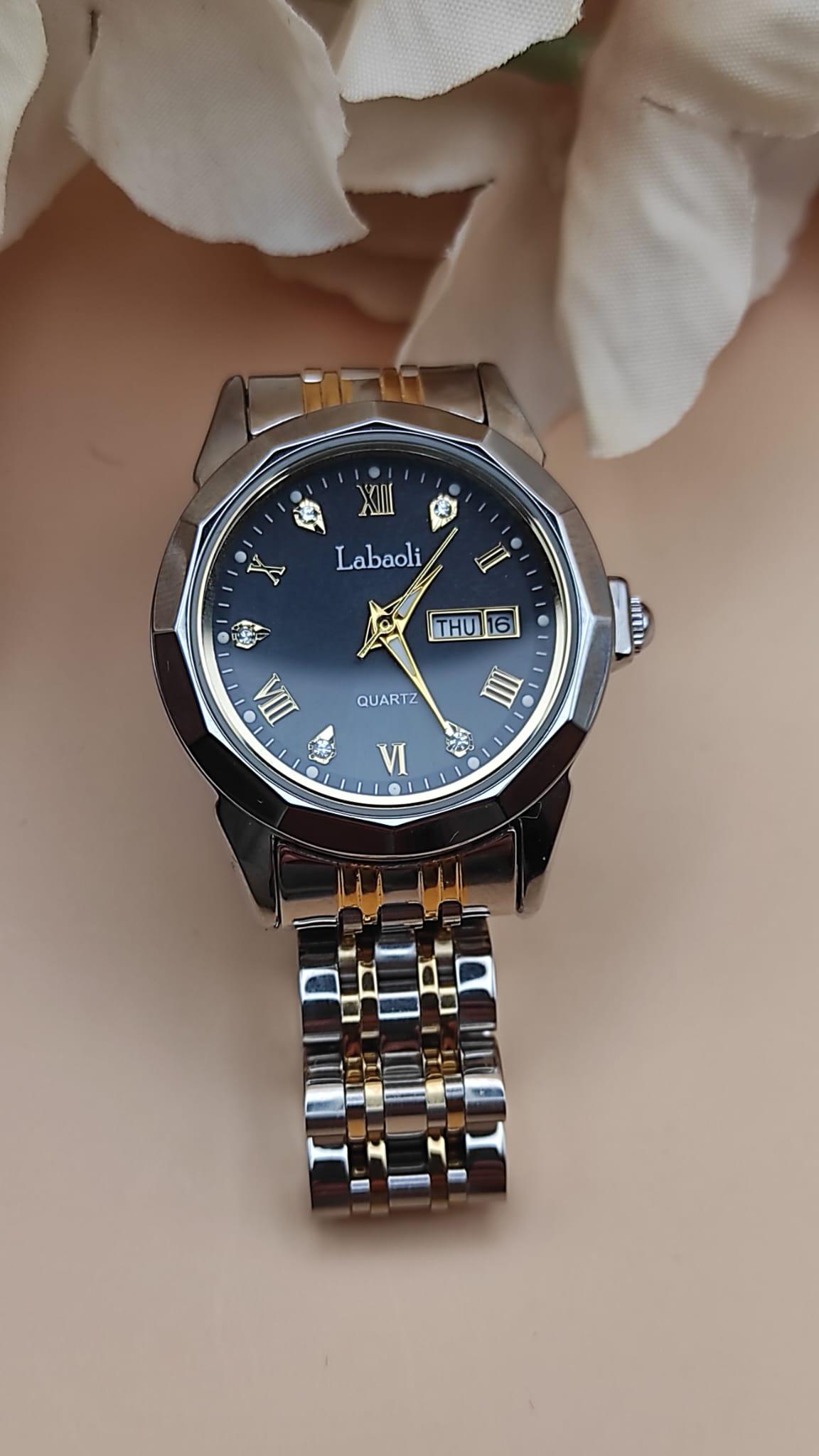 Montre Labaoli à cadran noir avec chiffres dorés stylisés, affichage jour et date à 3h, bracelet métallique bicolore argent et or, posée sur un fond floral beige.