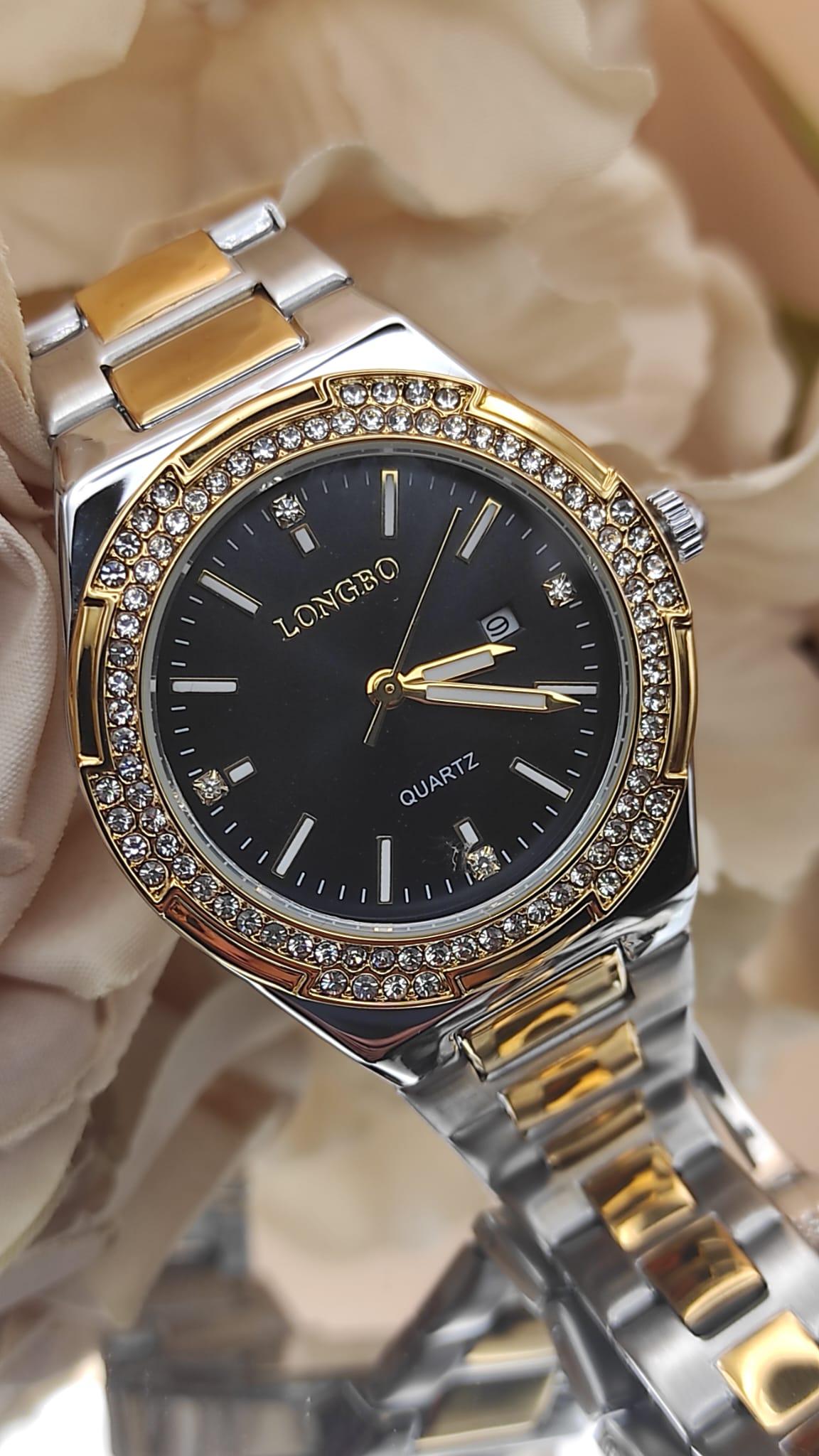 Montre LONGBO à cadran noir avec index dorés et pierres brillantes, lunette sertie de cristaux, bracelet métallique bicolore argent et or, posée sur un fond floral beige.