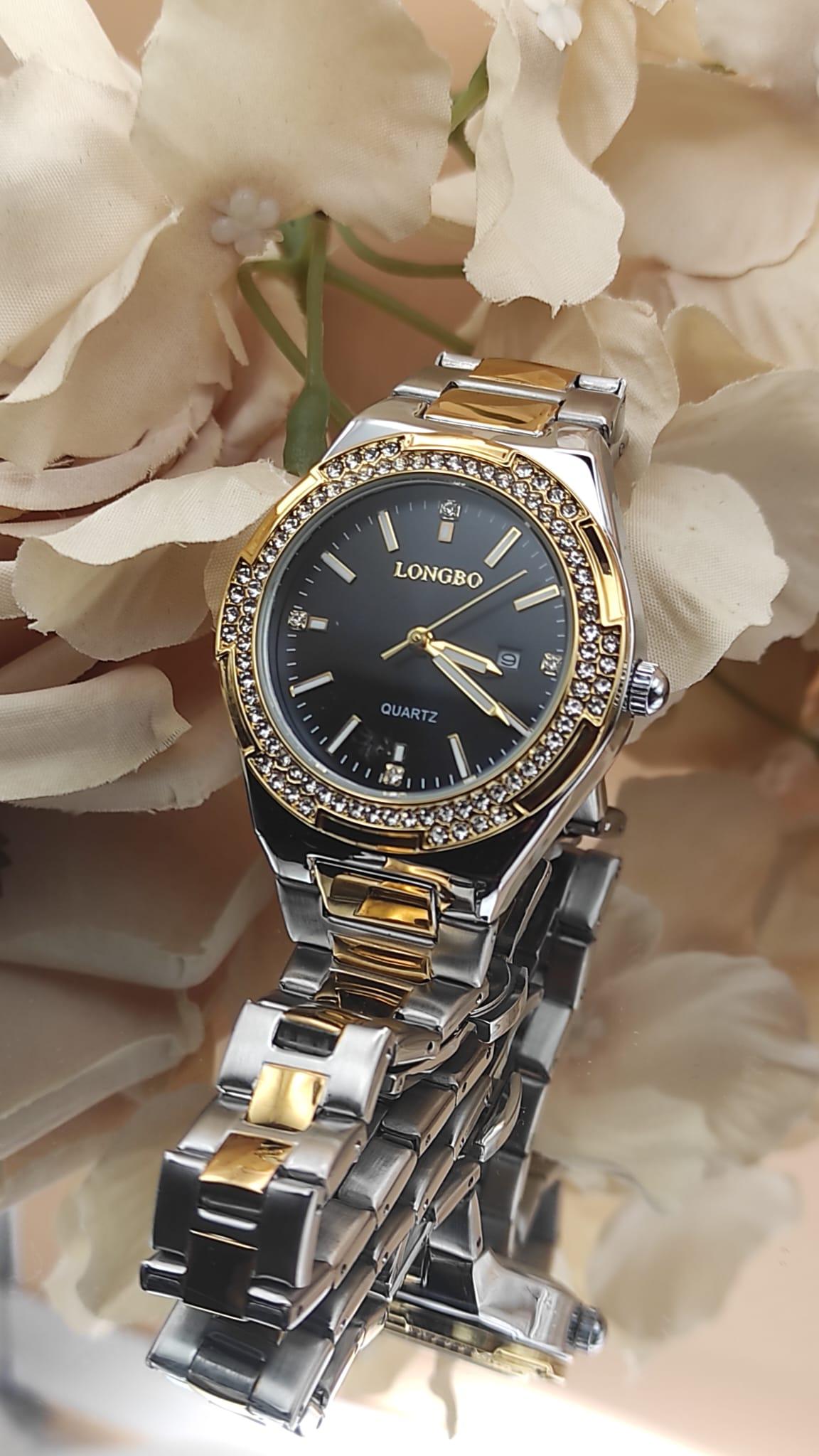 Montre LONGBO à cadran noir avec index dorés et pierres brillantes, lunette sertie de cristaux, bracelet métallique bicolore argent et or, posée sur un fond floral beige.