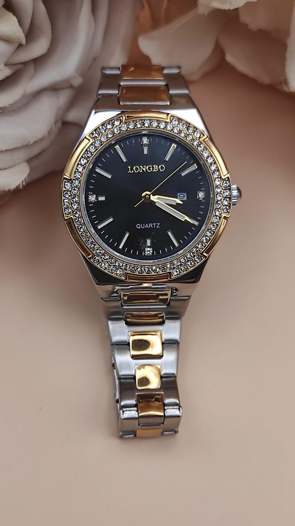 Montre LONGBO à cadran noir avec index dorés et pierres brillantes, lunette sertie de cristaux, bracelet métallique bicolore argent et or, posée sur un fond floral beige.
