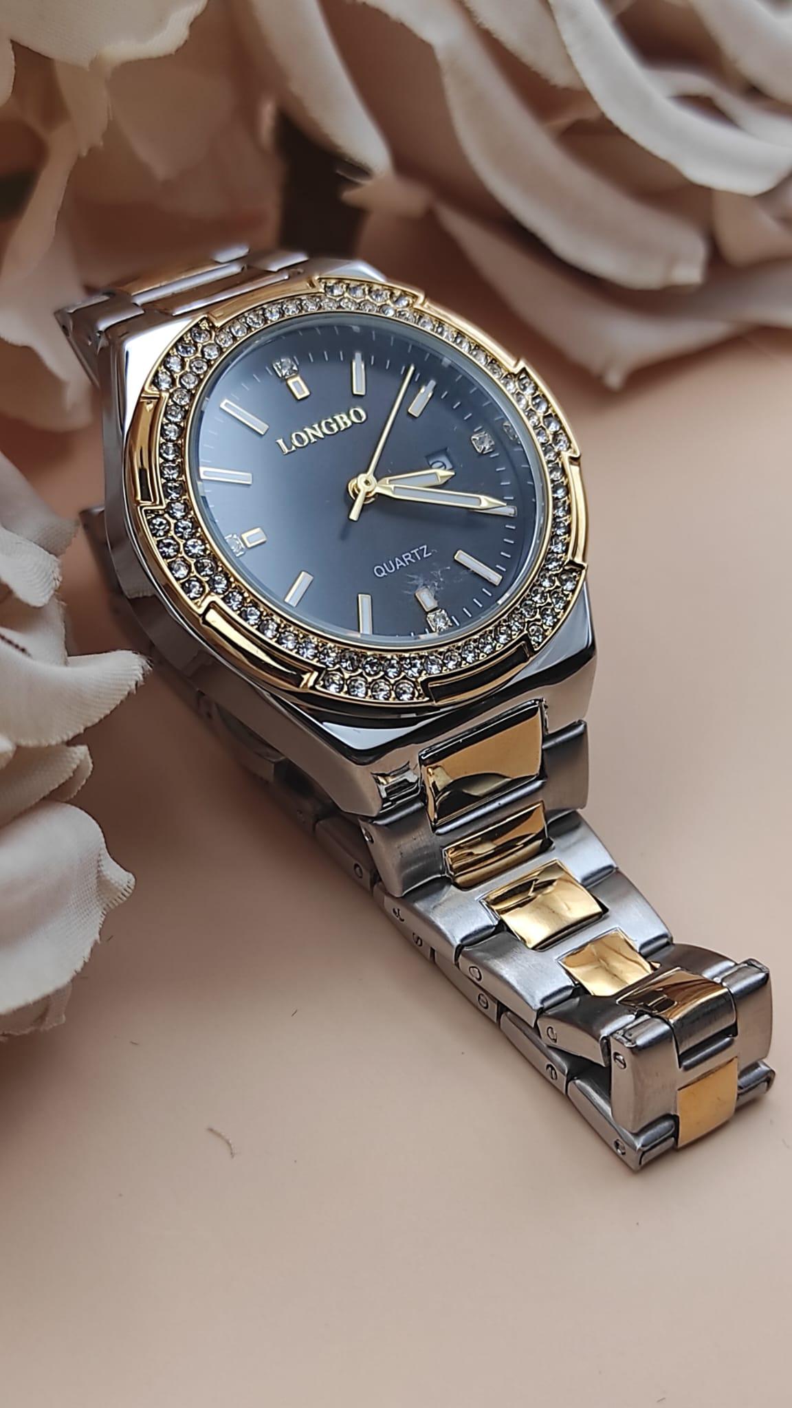 Montre LONGBO à cadran noir avec index dorés et pierres brillantes, lunette sertie de cristaux, bracelet métallique bicolore argent et or, posée sur un fond floral beige.